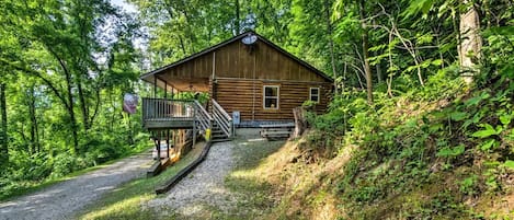 A wooden cabin with a porch and a staircase leading up to it, surrounded by dense forest.