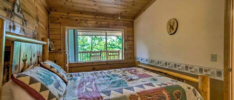 A wooden cabin bedroom with a large bed, a ceiling fan, a window with a view of greenery, and a wall clock.