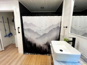 A bathroom with a shower curtain depicting mountains and birds, a white sink, and a wooden floor. - Modern Cabin in Pigeon Forge for a Romantic Glamping Rendezvous (Sevierville)