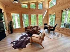 A cabin interior with a wooden floor, a sofa, a chair, a dining table, and large windows. - Modern Cabin in Pigeon Forge for a Romantic Glamping Rendezvous (Sevierville)