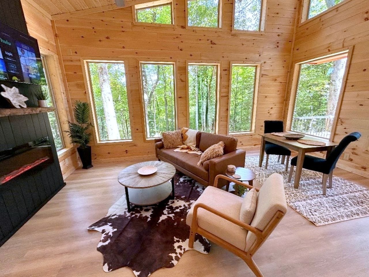 A cabin interior with a wooden floor, a sofa, a round coffee table, a dining table with chairs, and a TV mounted on the wall.