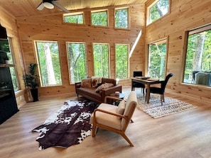 Ein Blockhaus-Interieur mit Holzboden, einer Couch, einem Sessel, einem Esstisch und großen Fenstern.