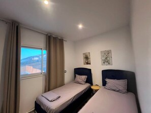 Ein Zimmer mit zwei Einzelbetten, einem fensterbelaubten Fenster, einem kleinen Tisch und zwei Wandbildern.