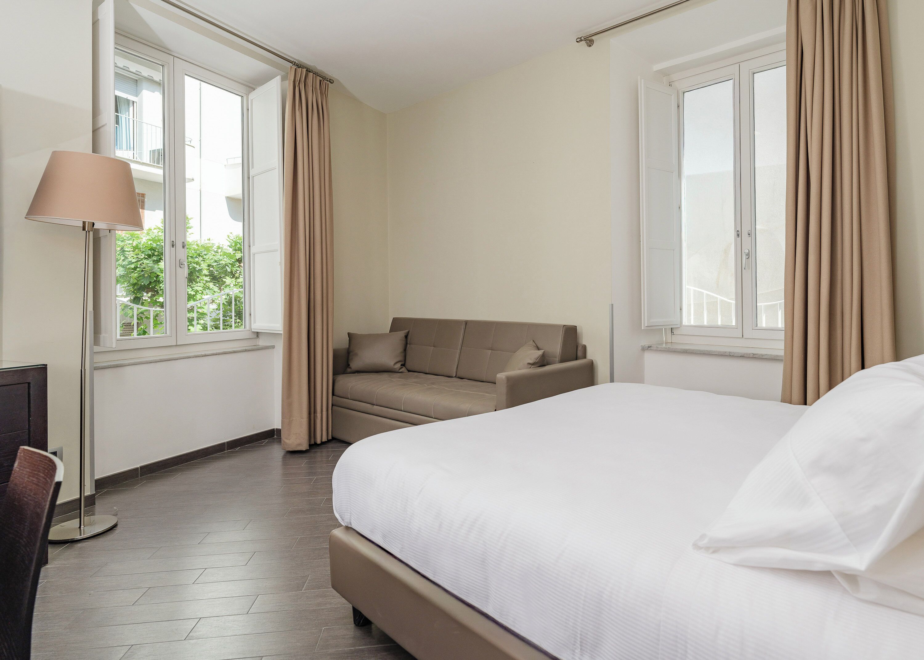 Una habitación de hotel con cama, sofá, lámpara y ventanas grandes.