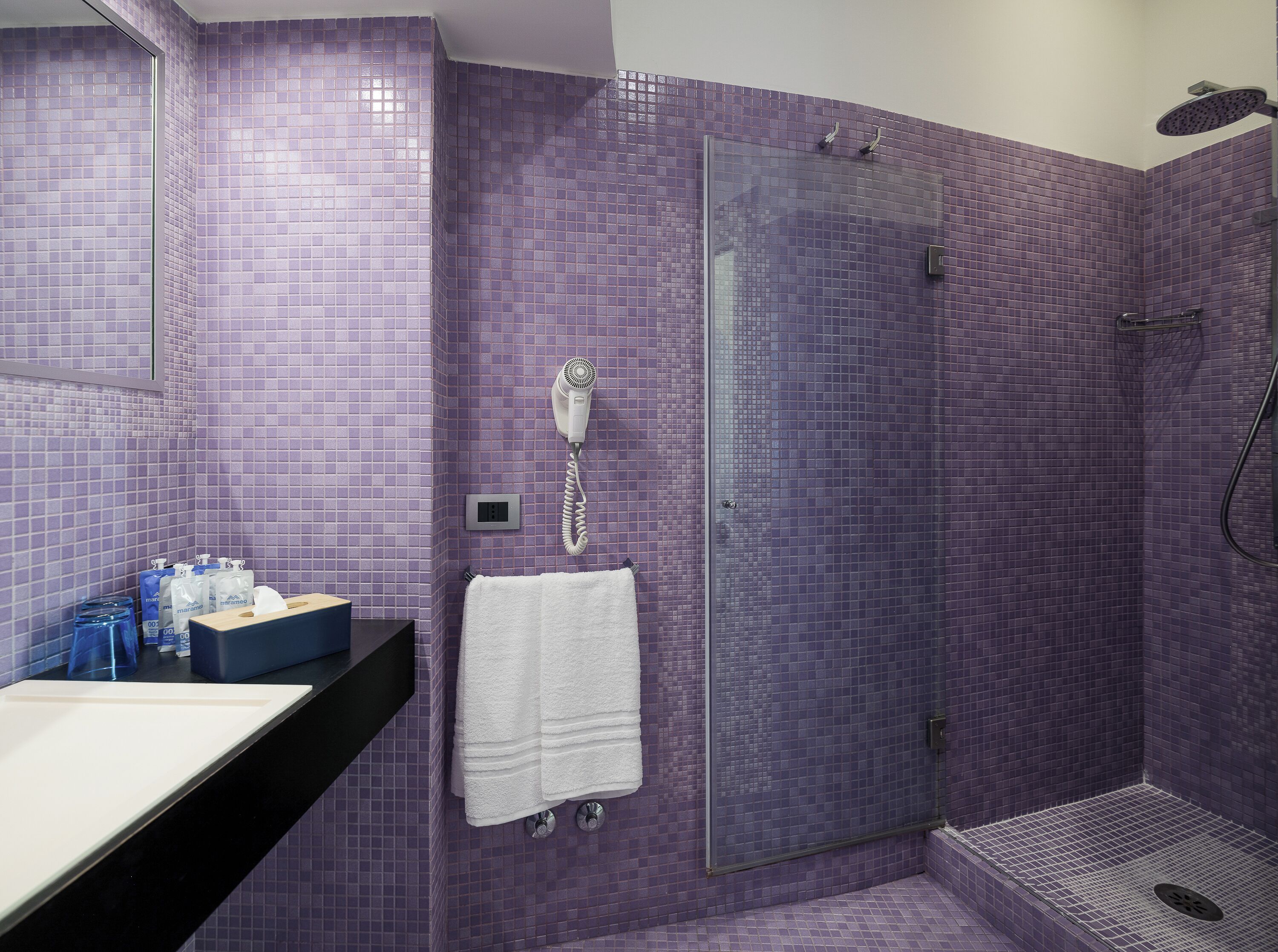 A bathroom with purple tiled walls, a glass shower enclosure, a white towel, and a hairdryer mounted on the wall.