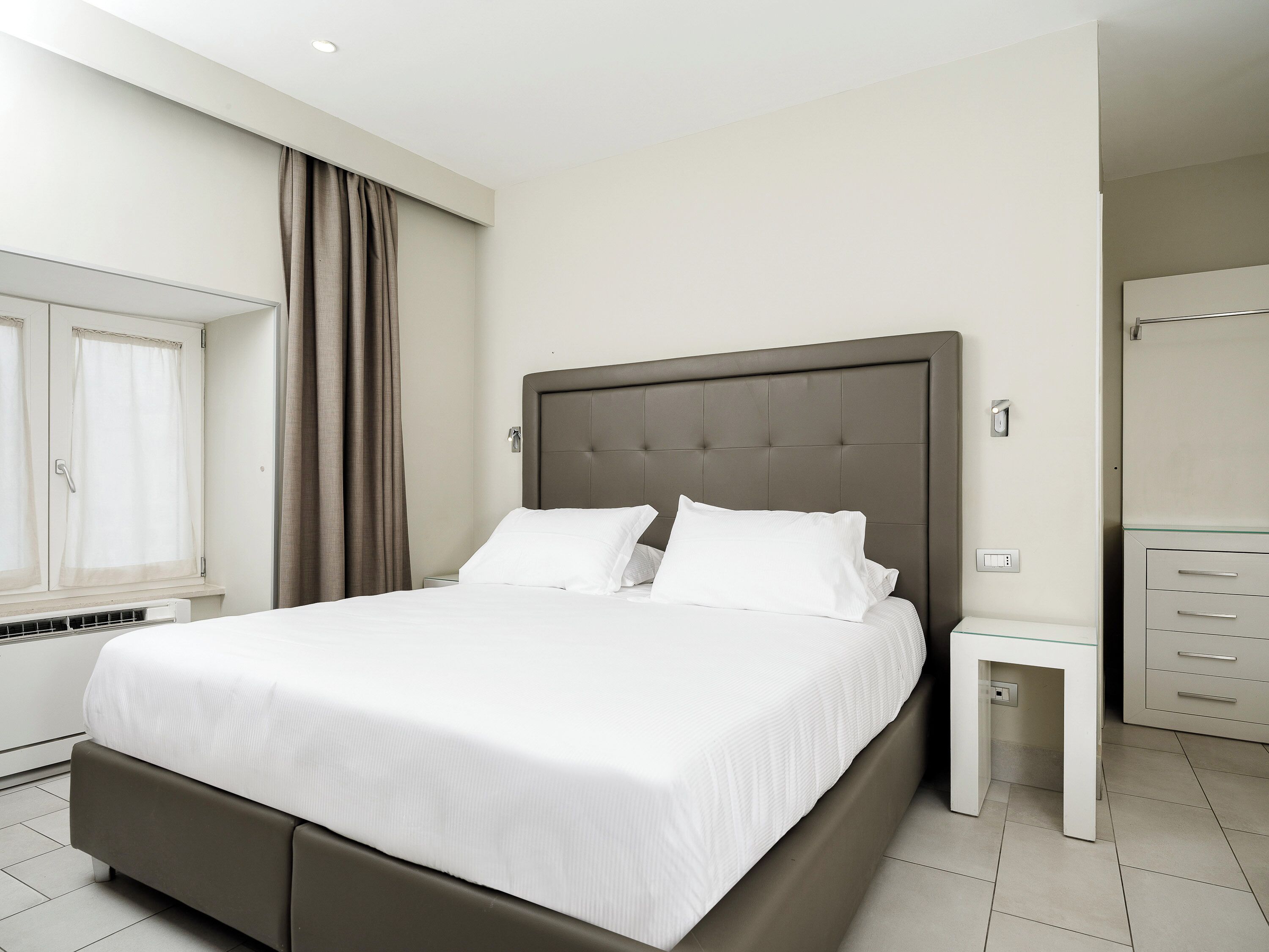 A modern hotel room with a large bed, a bedside table, a wardrobe, and a window with curtains.