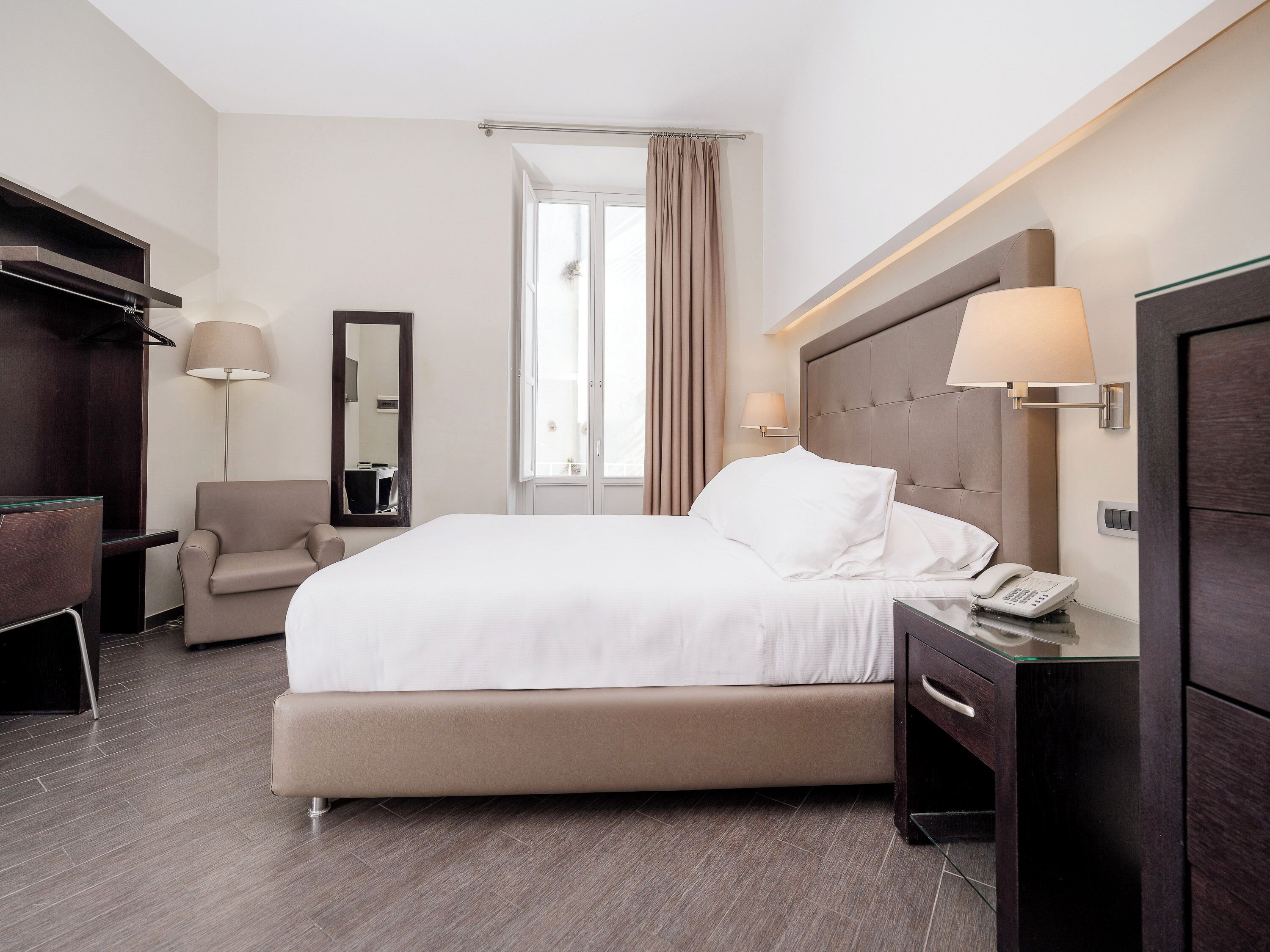 Una habitación de hotel con cama, una silla, escritorio con teléfono, espejo y televisor.