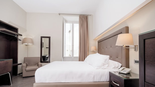 Une chambre d’hôtel avec un lit, une chaise, un bureau avec un téléphone, un miroir et une télévision.