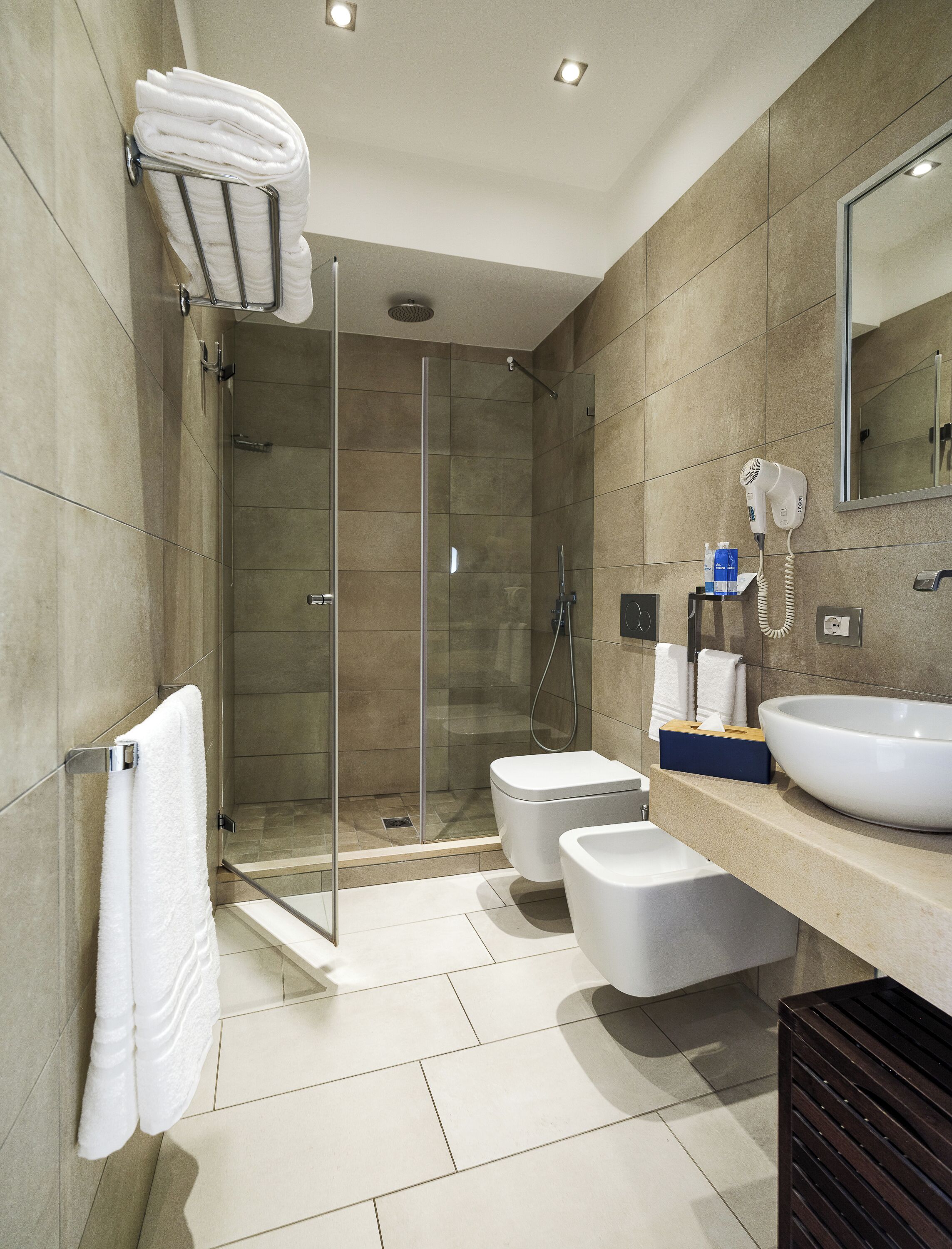 Un baño moderno con ducha envuelta en vidrio, inodoro, lavabo con una gran pileta y secador de pelo fijado a la pared.
