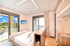 A modern bedroom with a large bed, a wooden bedside table, a TV, and a balcony with a glass door. - Minoas Sea Villa Heated Pool (Lake Kournas)