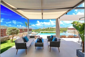 A covered outdoor seating area with a pool view, white sofas, and a table with flowers. - Minoas Sea Villa Heated Pool (Lake Kournas)