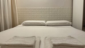 Een net opgemaakt bed met witte lakens en kussens, een gestructureerd hoofdbord in het wit, en twee opgevouwen handdoeken op het bed.