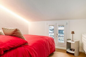 A bedroom with a red bed, a small bedside table with a lamp, and a window with a view of buildings. - A comfortable and welcoming 30 square meters studio apartment, perfect for three people, located on the fourth floor of a renovated building (no elevator).The property is situated in the heart of Modena (within the ZTL restricted traffic zone); it is an  (Modena)