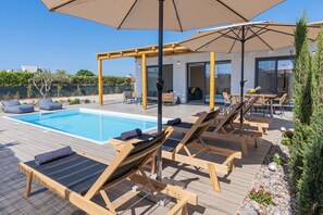Un espace aménagé au bord de la piscine, avec des chaises longues et des parasols, une pergola et une vue sur un bâtiment moderne.