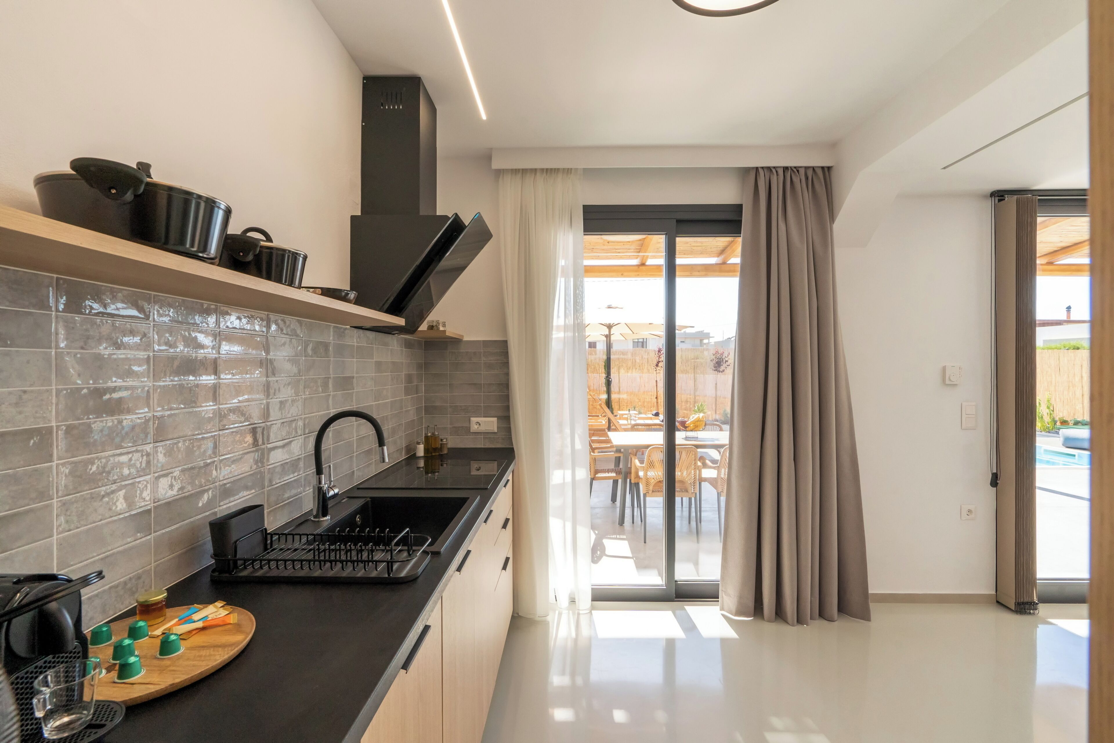 A modern kitchen with a black countertop, grey tiled backsplash, and a sliding glass door leading to an outdoor area.