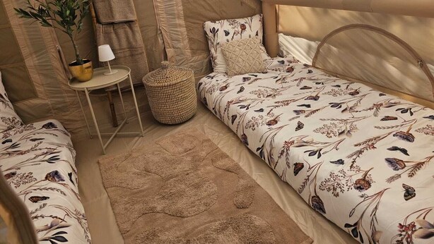 A cozy tent-style room with two beds, a small table with a lamp, a woven basket, and a potted plant.