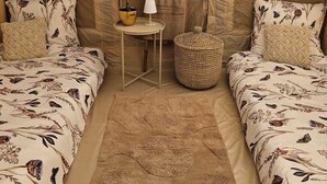 A tented room with two beds, a small table, a lamp, a potted plant, and a woven basket.