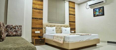 A bedroom with a bed, a nightstand, a sofa, a window with blinds, an air conditioning unit, and a painting on the wall.