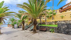 A paved walkway lined with palm trees, a stone wall, and a building with balconies. - Pueblo Canario Ocean Breeze Apartment (Playa de las Américas)