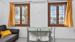 A room with a glass-top table, four chairs, a sofa with a yellow pillow, two windows with wooden frames and yellow curtains, and an air conditioning unit. - Pueblo Canario Ocean Breeze Apartment (Playa de las Américas)