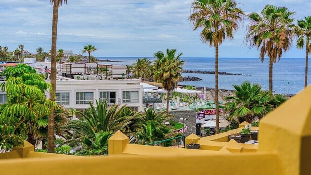 A coastal view with palm trees, a beach, and a building with a white facade. - Pueblo Canario Ocean Breeze Apartment (Playa de las Américas)