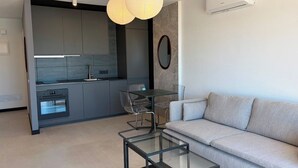 Et moderne køkken med indbygget ovn, et spiseområde med et glasbord og stole, samt en opholdsstue med sofa og sofabord.