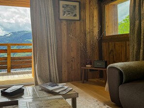 A room with a wooden interior, a view of a mountain landscape, a table with books, a chair, and a small shelf with a radio. - Nid du Mage: Breathtaking View of Megève (Megève)