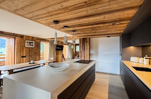 A modern kitchen with wooden beams, a large island, pendant lights, and a view of the outdoors. - Coeur de Mage: Stunning View of Megève (Megève)