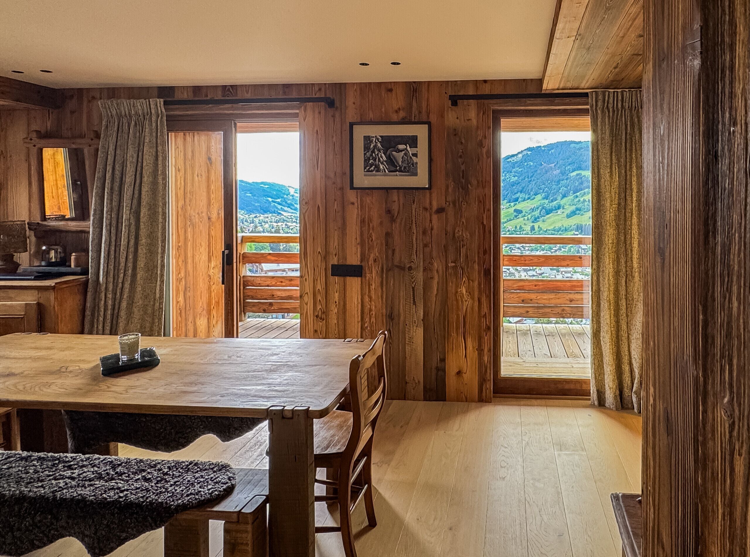 A wooden interior with a dining table, chairs, and a view of a mountainous landscape through large windows.