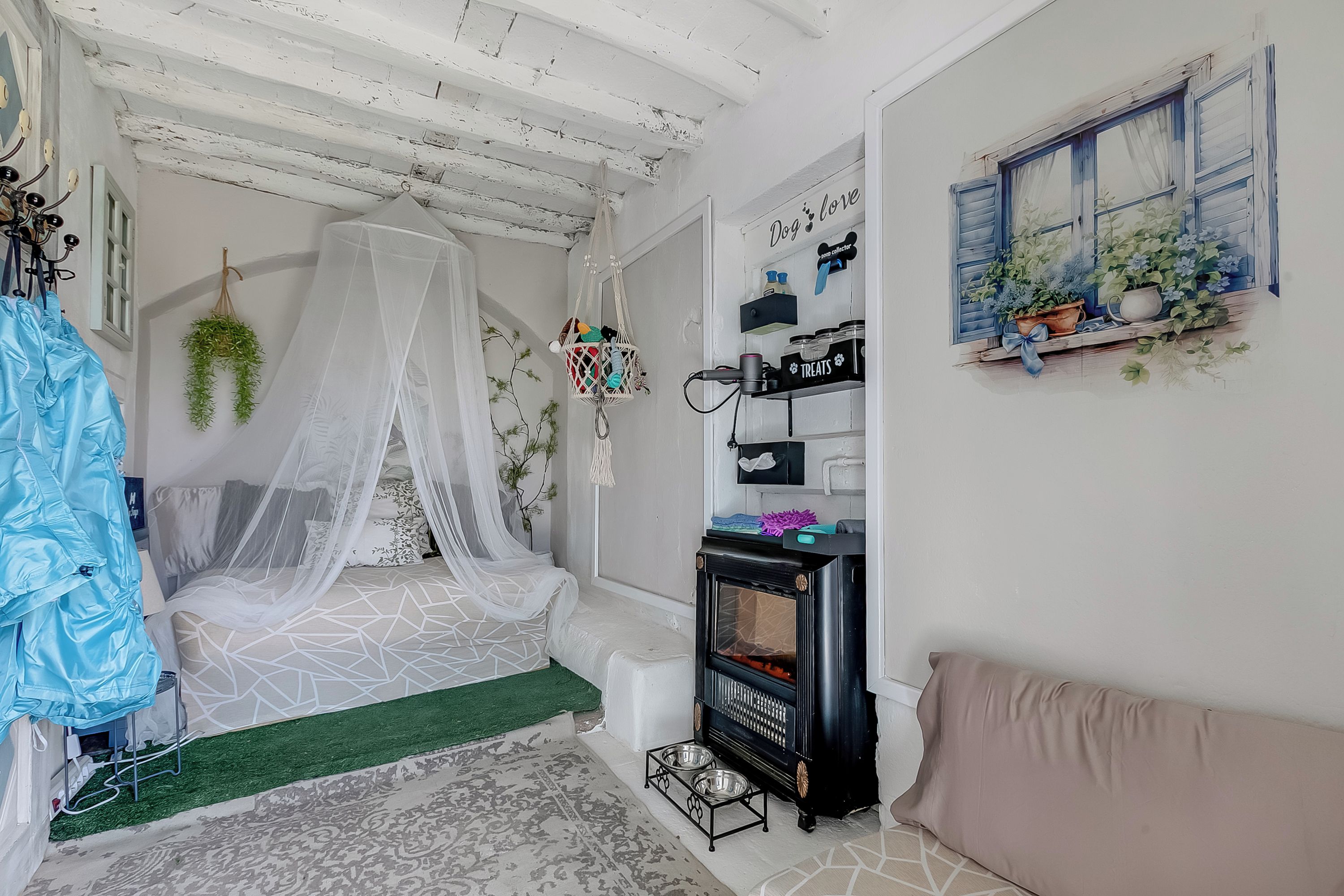 Une chambre à coucher avec un lit sous un baldaquin blanc, un petit radiateur noir, un panier pour chien, une étagère fixée au mur avec des plantes et une fenêtre avec des volets bleus.