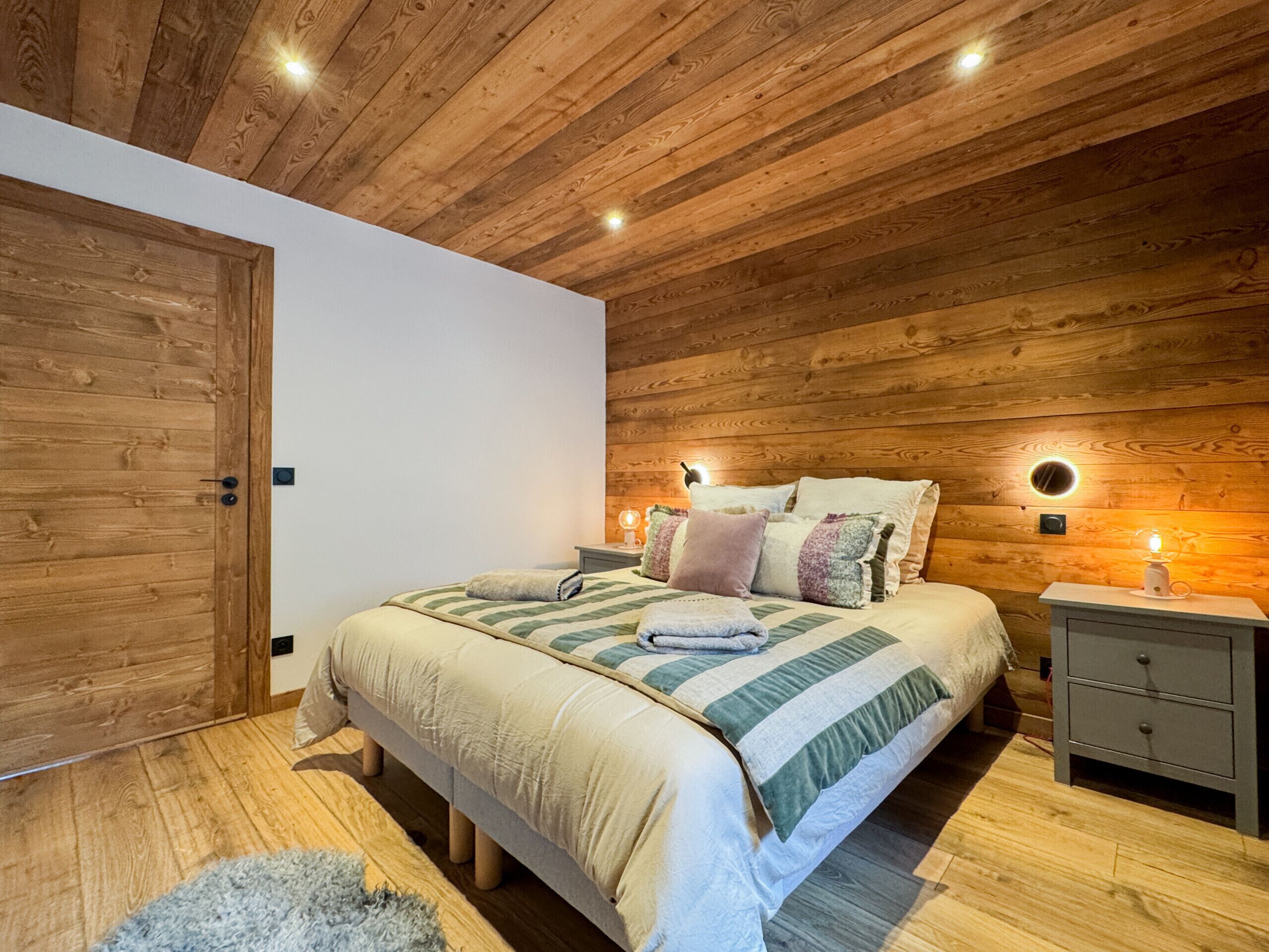 Een slaapkamer met een houten plafond, een bed met meerdere kussens, nachtkastjes met lampen, en een houten deur.