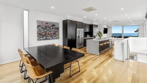 A modern kitchen with dark cabinetry, a central island, and a dining area with a long table and wooden chairs. - Ocean Views - just metres from the beach! (Ocean Grove)