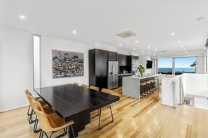 A modern kitchen with dark cabinetry, a central island, and a dining area with a long table and wooden chairs. - Ocean Views - just metres from the beach! (Ocean Grove)