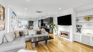 A modern living room with a sectional sofa, a flat-screen TV, a fireplace, and a kitchen in the background. - Ocean Views - just metres from the beach! (Ocean Grove)