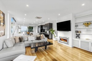 A modern living room with a sectional sofa, a flat-screen TV, a fireplace, and a kitchen in the background. - Ocean Views - just metres from the beach! (Ocean Grove)