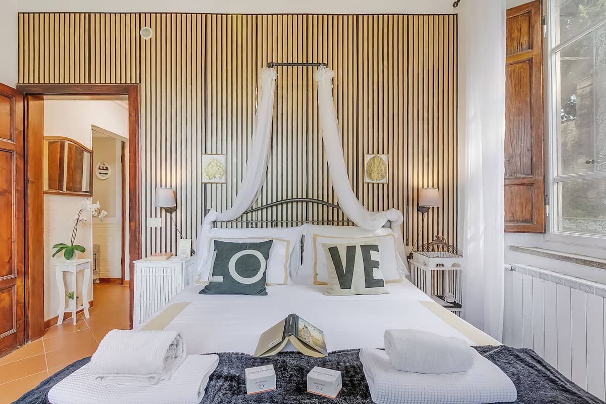 A neatly arranged bedroom with a canopy bed, a bedside table with a lamp, and a view of the outside through a window.