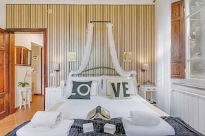 Ein ordentlich eingerichtetes Schlafzimmer mit einem Himmelsbett, einem Nachttisch mit Lampe und Blick ins Freie durch ein Fenster.