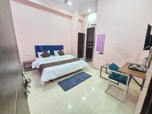 A bedroom with a bed, bedside table, desk, chair, and a door. - Hotel Delight (Jaipur)