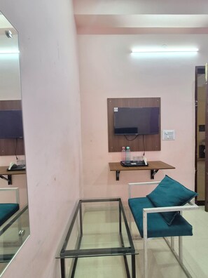 Una habitación con un televisor de pantalla plana montado en un panel de madera, una mesita con una bandeja que contiene un teléfono y botellas de agua, una mesita con superficie de vidrio y un cojín color turquesa sobre una estructura de metal blanco.