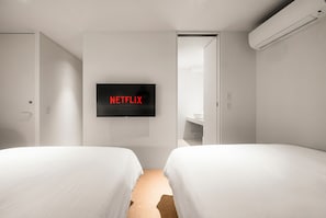 Habitació d'hotel moderna amb dos llits, un televisor fixat a la paret que mostra 'NETFLIX' i un bany a la vista.