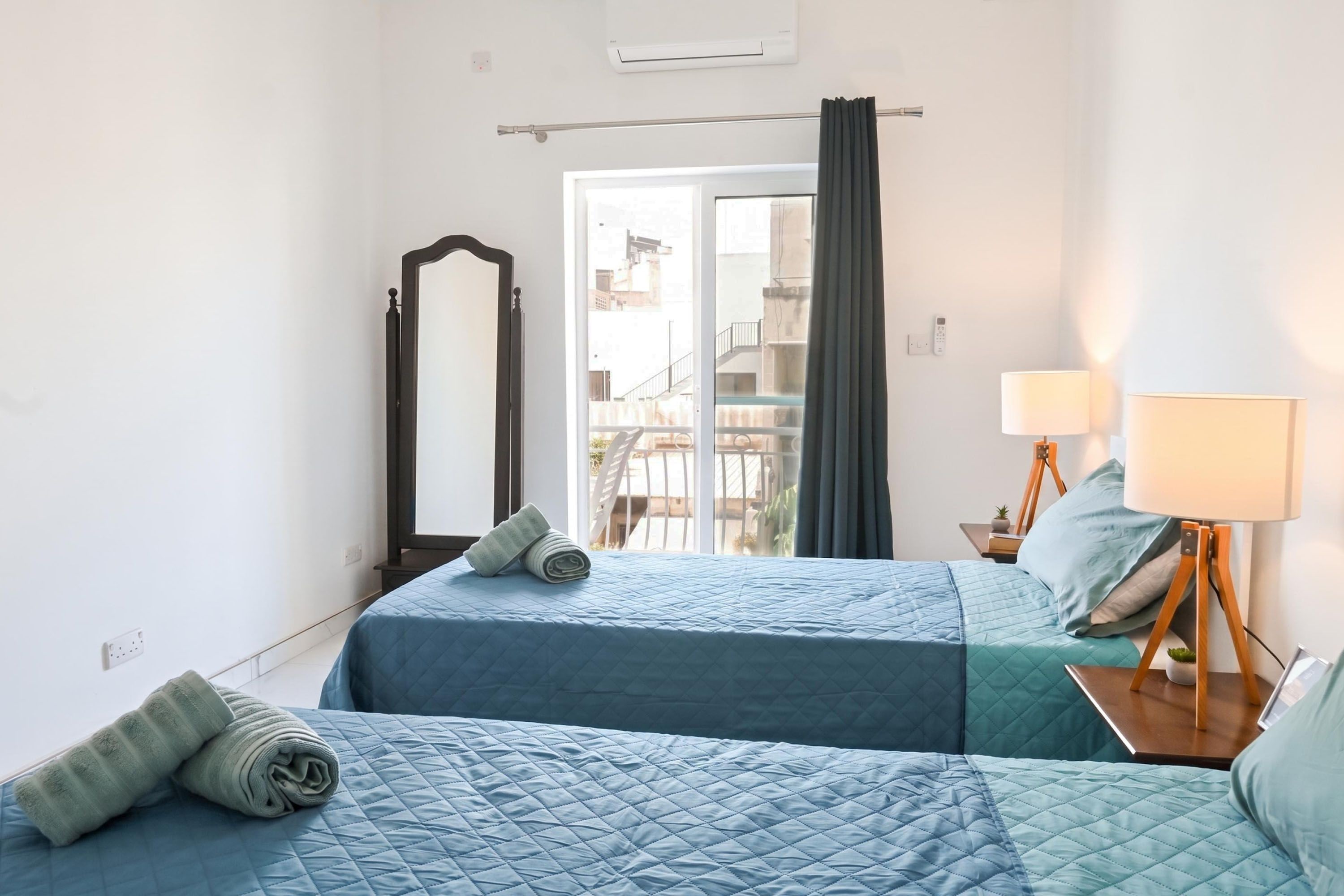 Habitación de hotel con dos camas, un espejo, una ventana con cortinas, una lámpara de noche y vistas al exterior.