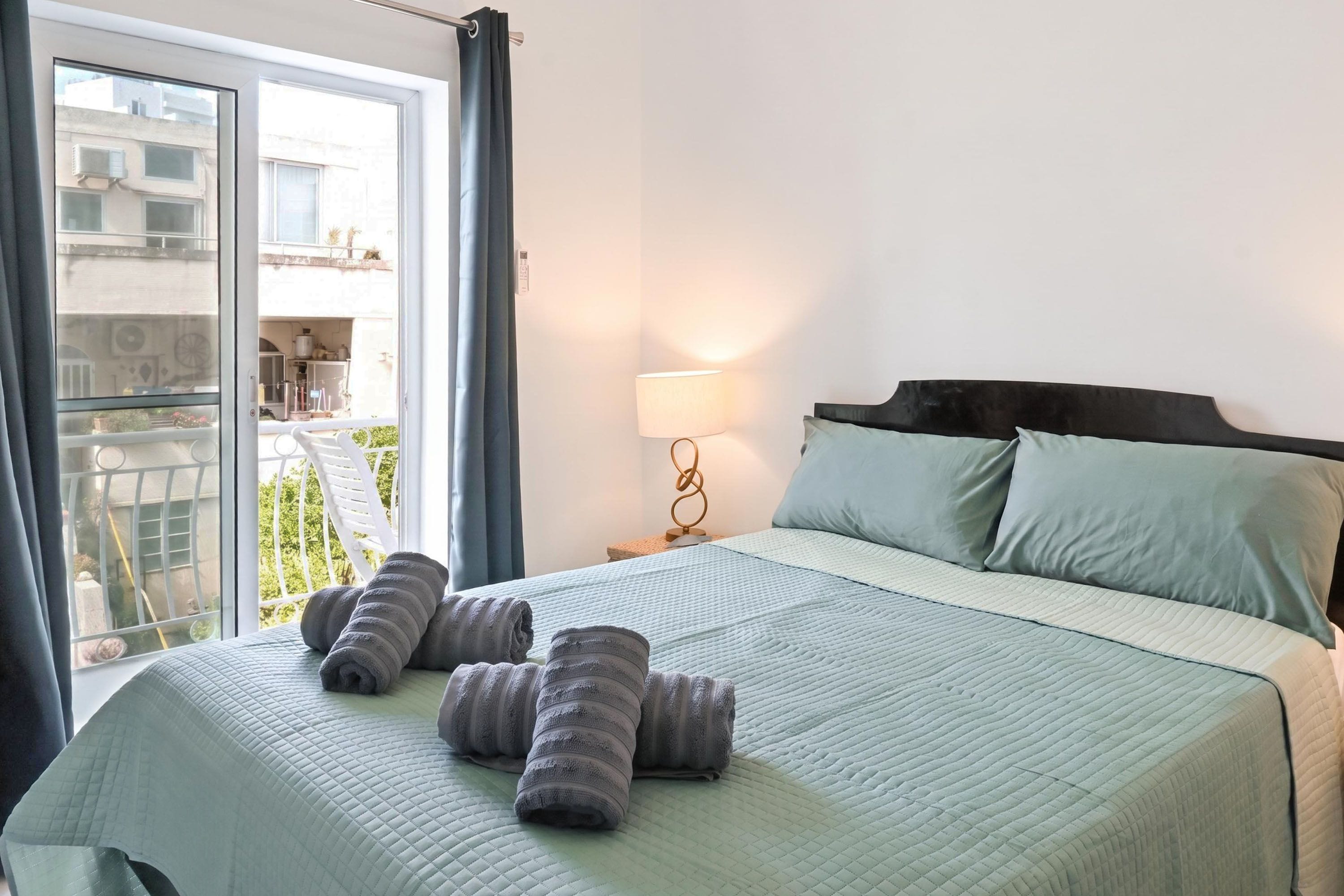 A hotel room with a bed, a nightstand, a lamp, and a view of the outside.
