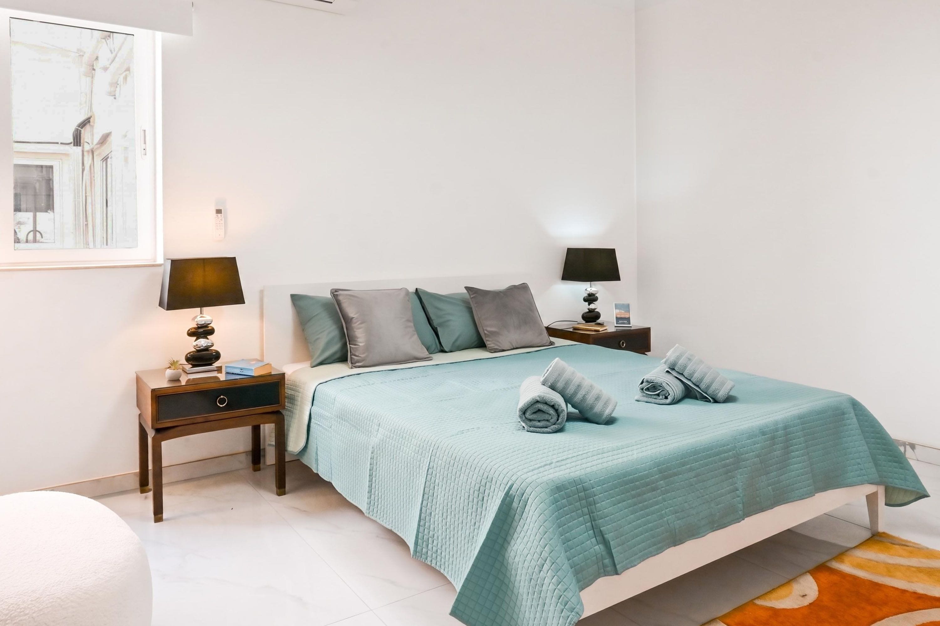 A bedroom with a bed, two bedside tables, a nightstand, a lamp, a painting, and a rolled towel on the bed.