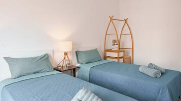 Una habitación con dos camas, cada una con colcha azul acolchada y almohadas a juego. Se observa una estantería de escalera de madera, una mesita de noche con lámpara y una pequeña cesta.