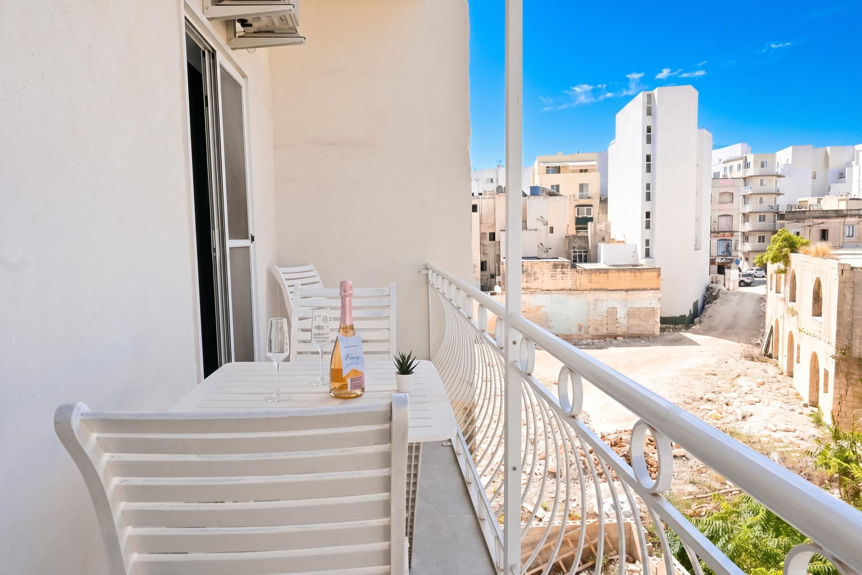 Ein Balkon mit einem weißen Tisch, einer Champagnerflasche und einem Glas. Der Balkon bietet Blick auf eine Stadtlandschaft mit Gebäuden und blauem Himmel.