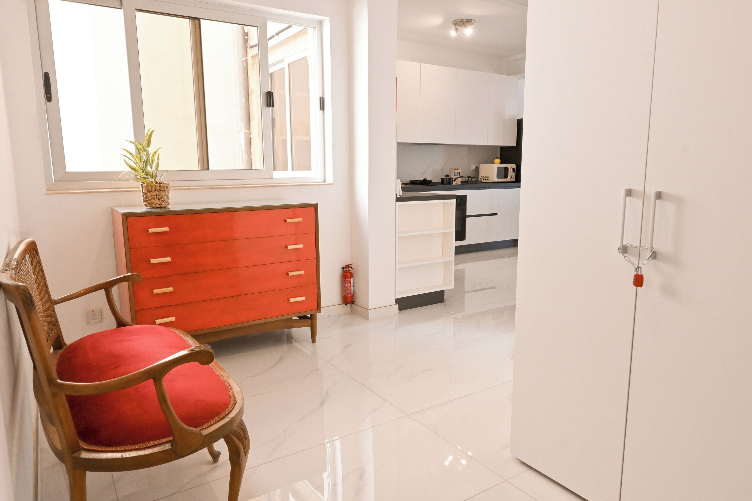 Ein Zimmer mit einem Holzstuhl, einer rot gepolsterten Sitzgelegenheit, einem weißen Kleiderschrank und einer Küchenzeile mit weißen Schränken und einer Mikrowelle.