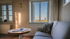 Ein Wohnzimmer mit einer Couch, einem runden Tisch mit Unterlage, einem Fenster mit Vorhängen und Blick auf den Strand.
