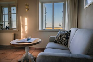 A living room with a sofa, a round table with a placemat, a window with curtains, and a view of the beach. - Holiday Home "Haus Nordmarsch" with Sea View, Private Terrace and Wi-Fi (Langeneß)