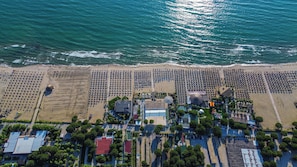 An aerial view of a beach resort with rows of sunbeds, a central building with a blue roof, and a clear view of the ocean.
