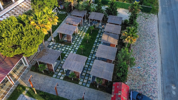 An aerial view of a resort with thatched-roof huts arranged in a grid pattern, surrounded by lush greenery and a paved walkway.